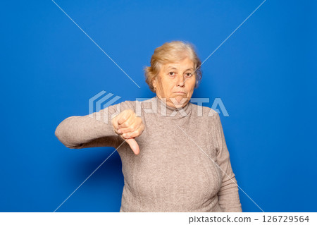 Caucasian woman in her 70s wearing a dark beige sweater with her thumb down in a sign of disgust and disapproval, isolated on blue studio background. 126729564