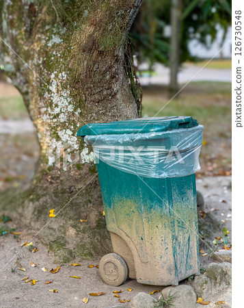 Trash bin with dirt in natural outdoor setting beside tree, showing urban waste and environmental decay 126730548