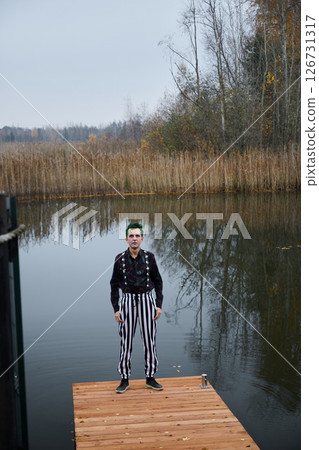 An Eerie Clown Standing on a Dock by the Water, Eliciting a Sense of Mystery and Suspense An Eerie Clown Standing on a Dock by the Water, Eliciting a Sense of Mystery and Suspense 126731317