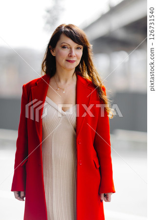 Elegant businesswoman walking in urban setting wearing red coat and beige dress 126731540