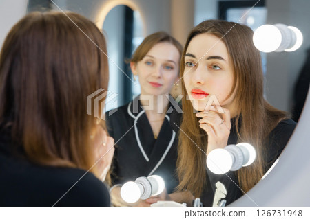 Woman applying makeup looking at mirror with beautician assisting her 126731948