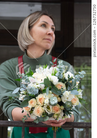 Close-up of a female florist in a green sweater and red suspenders holding a bouquet of cream roses, white alstroemeria, and eucalyptus Close-up of a female florist in a green sweater and red suspenders holding a bouquet of cream roses, white alstroemeria, and eucalyptus 126732750