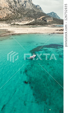 A small boat sailing in the azure water of the sea near a rocky beach in Balos Lagoon 126732835