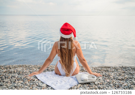 A woman wearing a red Santa hat sits on a beach towel. She is wearing a bikini and has a laptop on her lap. A woman wearing a red Santa hat sits on a beach towel. She is wearing a bikini and has a laptop on her lap. 126733186