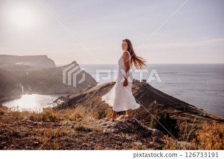 A woman in a white dress stands on a hill overlooking the ocean. The scene is serene and peaceful, with the woman's dress billowing in the wind. The combination of the ocean. A woman in a white dress stands on a hill overlooking the ocean. The scene is serene and peaceful, with the woman's dress billowing in the wind. The combination of the ocean. 126733191