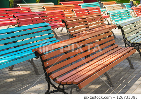 Row of multi colored benches. Colorful wooden benches arranged in rows outdoors, creating lively playful atmosphere in urban park. Concept of color harmony, outdoor relaxation. City urban background Row of multi colored benches. Colorful wooden benches arranged in rows outdoors, creating lively playful atmosphere in urban park. Concept of color harmony, outdoor relaxation. City urban background 126733383