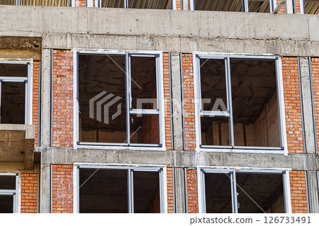 The exterior of a brick multi-storey building under construction during the installation of window frames with preliminary thermal insulation works. There are no people, no workers The exterior of a brick multi-storey building under construction during the installation of window frames with preliminary thermal insulation works. There are no people, no workers 126733491