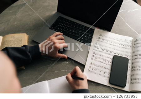 High-angle view of unrecognizable composer male creating music notes on paper while using laptop, surrounded by sheet music, book, smartphone on table in cozy home workspace. 126733953