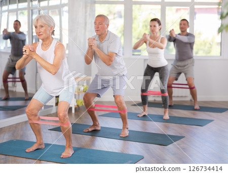 Elderly woman during Pilates strength training performs squats with elastic band on legs and shins Elderly woman during Pilates strength training performs squats with elastic band on legs and shins 126734414