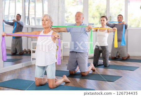 Elderly woman kneeling performs exercises with wide elastic band during Pilates workout. 126734435