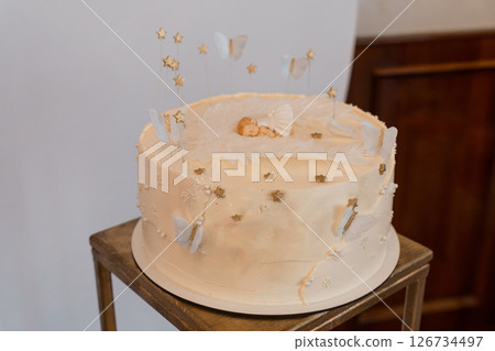 Beautifully Decorated Baby Shower Cake with a Sleeping Baby Topper and Golden Accents on a Wooden Stand, Featuring Stars and Butterflies 126734497