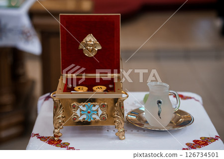 A detailed view of a red ornate box with a golden cross on top, positioned on a table covered with a red embroidered cloth and liturgical objects 126734501