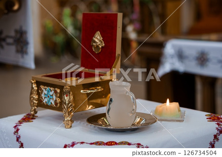 A solemn religious scene depicting a red ornate box open on a table alongside a white ceramic cup and golden receptacle, illuminated by a candle glow inside a church 126734504