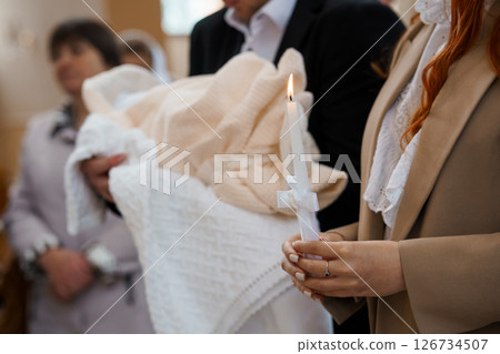 A blurred image capturing a Christian baptism ceremony, featuring a baby wrapped in a white blanket held by a woman, with family members surrounding them holding lit candles 126734507