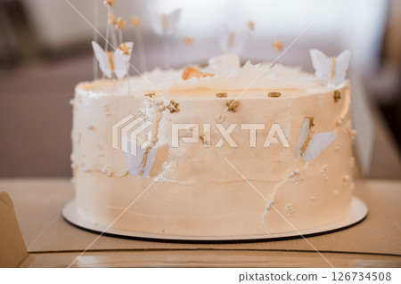 A beautifully decorated light peach frosted cake adorned with delicate white butterflies and golden stars, set on a wooden surface, creating a whimsical and elegant dessert presentation 126734508