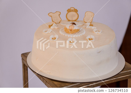 A beautifully decorated round vanilla cake adorned with delicate white flowers and golden baby items, symbolizing a newborn celebration, sits on a wooden stand A beautifully decorated round vanilla cake adorned with delicate white flowers and golden baby items, symbolizing a newborn celebration, sits on a wooden stand 126734510