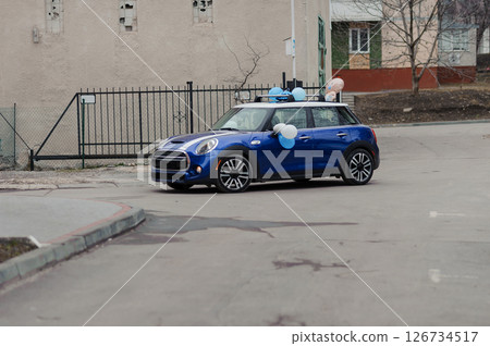 A blue Mini Cooper parked on a street with balloons attached to its roof, celebrating a baby boy, in front of a beige building and wrought iron fence 126734517