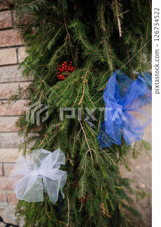 Festive Holiday Wreath Decoration with Evergreen Branches, Red Berries, and Blue and White Ribbons Against Brick Wall Background, Winter Season 126734522