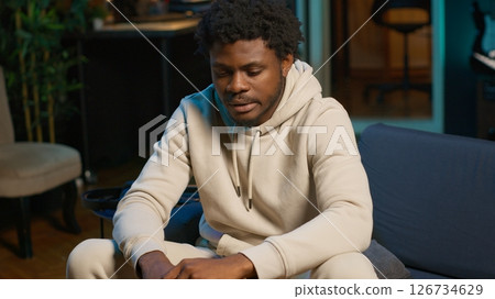 African american male remote worker sitting at home on couch late at night, overthinking about problems. Young man struggling to finish work project before deadline, finding solutions. Camera B. 126734629