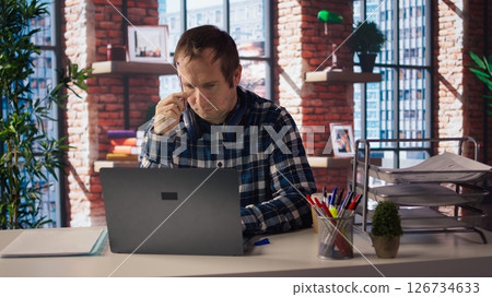 Stressed man in home office scratching head with pen, upset after having to answer too many emails on laptop. Fatigued teleworker feeling overworked by too many project tasks, camera A 126734633