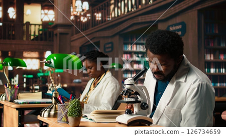 Male student researching viruses during a microscope analysis and academic reading in a classic library. Med school pupil reflects dedication to biology and exam preparations. Camera A. Male student researching viruses during a microscope analysis and academic reading in a classic library. Med school pupil reflects dedication to biology and exam preparations. Camera A. 126734659