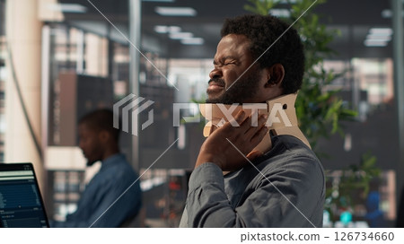 Hurt employee with a cervical collar working on a new business project, struggling to sit at the desk with his neck fracture. African american company associate tries to solve tasks. Camera A. 126734660