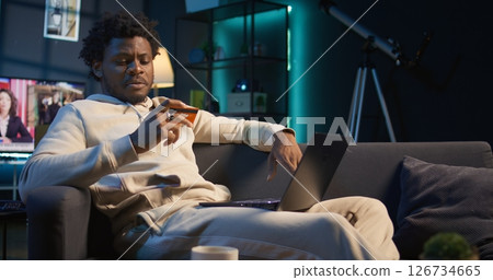 Smiling african american man entering card digits to confirm a money transfer, setting up the payment for online shopping on his pc. Happy person feeling good after placing orders. Camera B. 126734665
