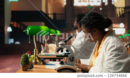 Dedicated female student working with a microscope to prepare for microbiology lessons in a library, focusing on microorganisms and chemical processes. Notes for her academic project. Camera A. 126734666