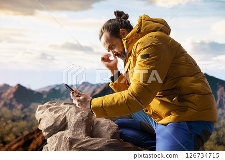 Young trekker being distracted by his phone on top of the mountain, dealing with technology addiction and always connected to the internet. Explorer enjoying scenic landscape view. Young trekker being distracted by his phone on top of the mountain, dealing with technology addiction and always connected to the internet. Explorer enjoying scenic landscape view. 126734715