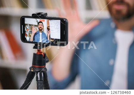Cropped photo of a phone screen recording man explaining some language lesson. Male tutor sitting at table and recording a video of himself on a phone. Tutoring remote, modern education at home Cropped photo of a phone screen recording man explaining some language lesson. Male tutor sitting at table and recording a video of himself on a phone. Tutoring remote, modern education at home 126738460
