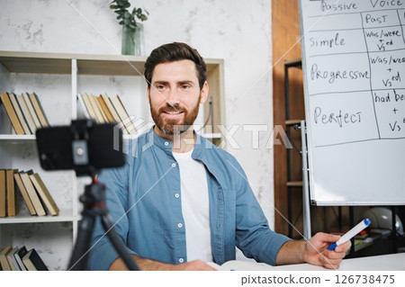 Male tutor sitting at table and recording a video of himself on a phone. Man recording video lessons. Tutoring remote, modern education at home, online lesson, study language concept. 126738475
