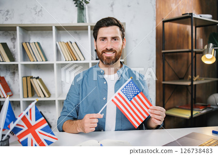 Excited male tutor sitting at table with flag of USA looking and smiling at camera, copy space. Man teaching foreign language online at home. Tutoring remote, modern education at home, online lesson 126738483