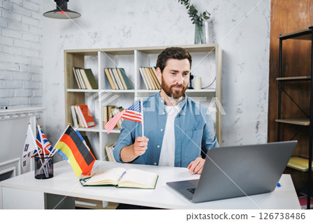 Male tutor sitting at table with flags of different countries looking at laptop. Man teaching foreign language online at home. Tutoring remote, modern education at home, online lesson, study language Male tutor sitting at table with flags of different countries looking at laptop. Man teaching foreign language online at home. Tutoring remote, modern education at home, online lesson, study language 126738486