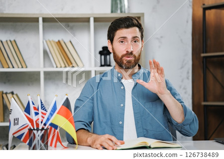 Male tutor sitting at table with flags of different countries looking at camera and explaining something. Man teaching foreign language online at home. Tutoring remote, modern education at home 126738489