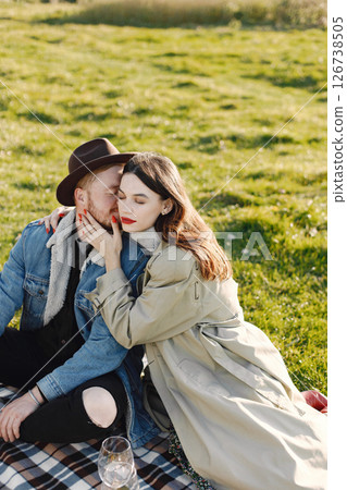 Man and woman in fashion clothes sitting on a nature on a picnic rug. Man wearing jacket and a black hat and woman coat. Woman hugging a man. 126738505