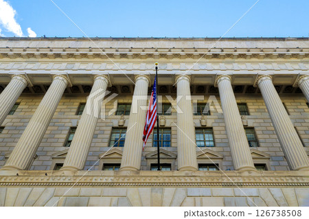 Tall columns support facade of government building, showcasing American flag under clear blue sky. 126738508