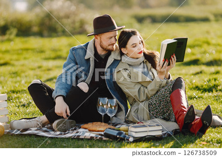 Man and woman in fashion clothes sitting on a nature on a picnic rug. Man wearing jacket and a black hat and woman skirt and red boots. Woman reding a book. 126738509