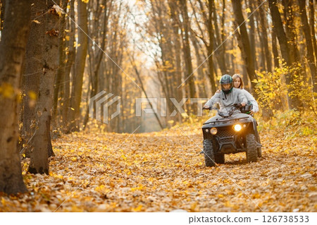 A young caucasian man and woman riding an ATV quad bike in autumn forest. Couple maneuvering off-road ATV. Couple wearing grey sportive costumes. 126738533