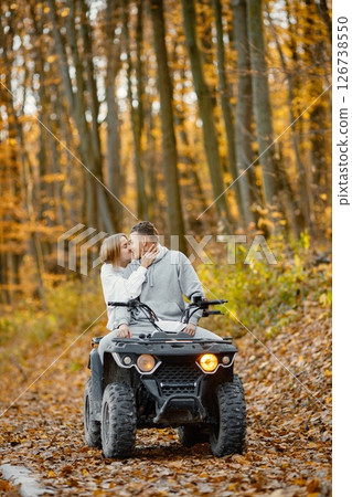 A young caucasian man and woman riding an ATV quad bike in autumn forest. Lovely couple kissing. Couple wearing grey sportive costumes. 126738550