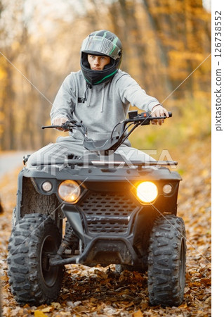 A young caucasian man in helmet riding an ATV quad bike in autumn forest. Boy maneuvering off-road ATV. Man wearing grey sportive costume. A young caucasian man in helmet riding an ATV quad bike in autumn forest. Boy maneuvering off-road ATV. Man wearing grey sportive costume. 126738552