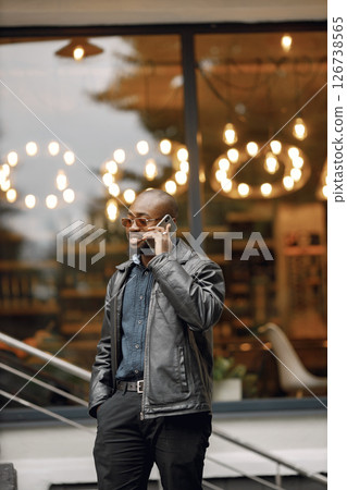 Black man standing on a street in a city. Man wearing sunglasses and a leather jacket. Business man using a phone. Black man standing on a street in a city. Man wearing sunglasses and a leather jacket. Business man using a phone. 126738565