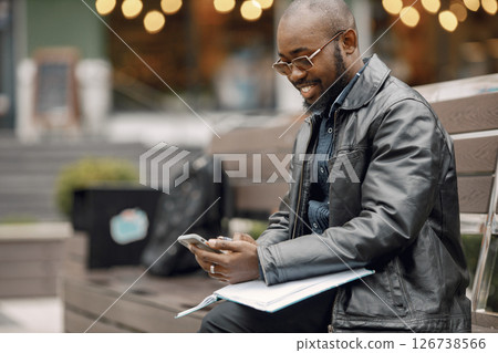 Black man sitting on a bench in a city. Man wearing sunglasses and a leather jacket. Business man has a papers and using a phone. Black man sitting on a bench in a city. Man wearing sunglasses and a leather jacket. Business man has a papers and using a phone. 126738566