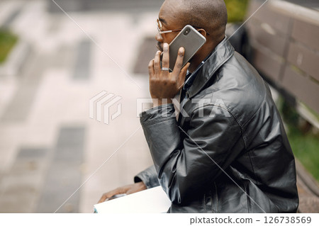 Black man sitting on a bench in a city. Man wearing sunglasses and a leather jacket. Business man has a papers and using a phone. 126738569