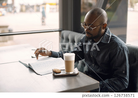 Black man sitting in a coffee shop and drinking a latte. Man wearing sunglasses and grey shirt. Business man working in a cafe. 126738571