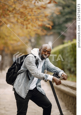 Black man standing on a street with electric scooter. Man wearing white hoodie and black jeans. Man with black backpack posing for a photo. 126738573