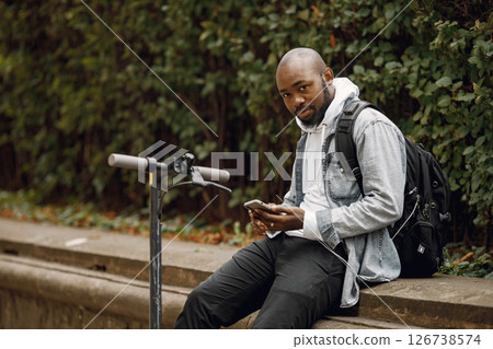 Black man sitting on a street with electric scooter. Man wearing white hoodie and black jeans. Man with black backpack using a phone. 126738574