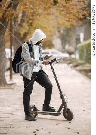 Black man standing on a street with electric scooter. Man wearing white hoodie and black jeans. Man with black backpack using a phone. 126738575