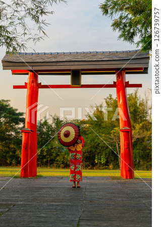 Back view of a young woman wearing a Japanese Yukata and holding a paper umbrella at Torii traditional Japanese gate. 126739757