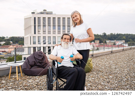 Elderly woman assists wheelchair-bound husband with exercise using dumbbells outdoors, promoting recovery and wellness. 126742497