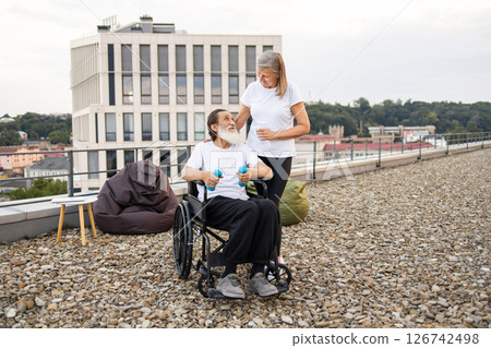 Elderly couple exercising together outdoors. Senior woman supporting her husband seated in wheelchair during physical activity focusing on recovery and strength. 126742498